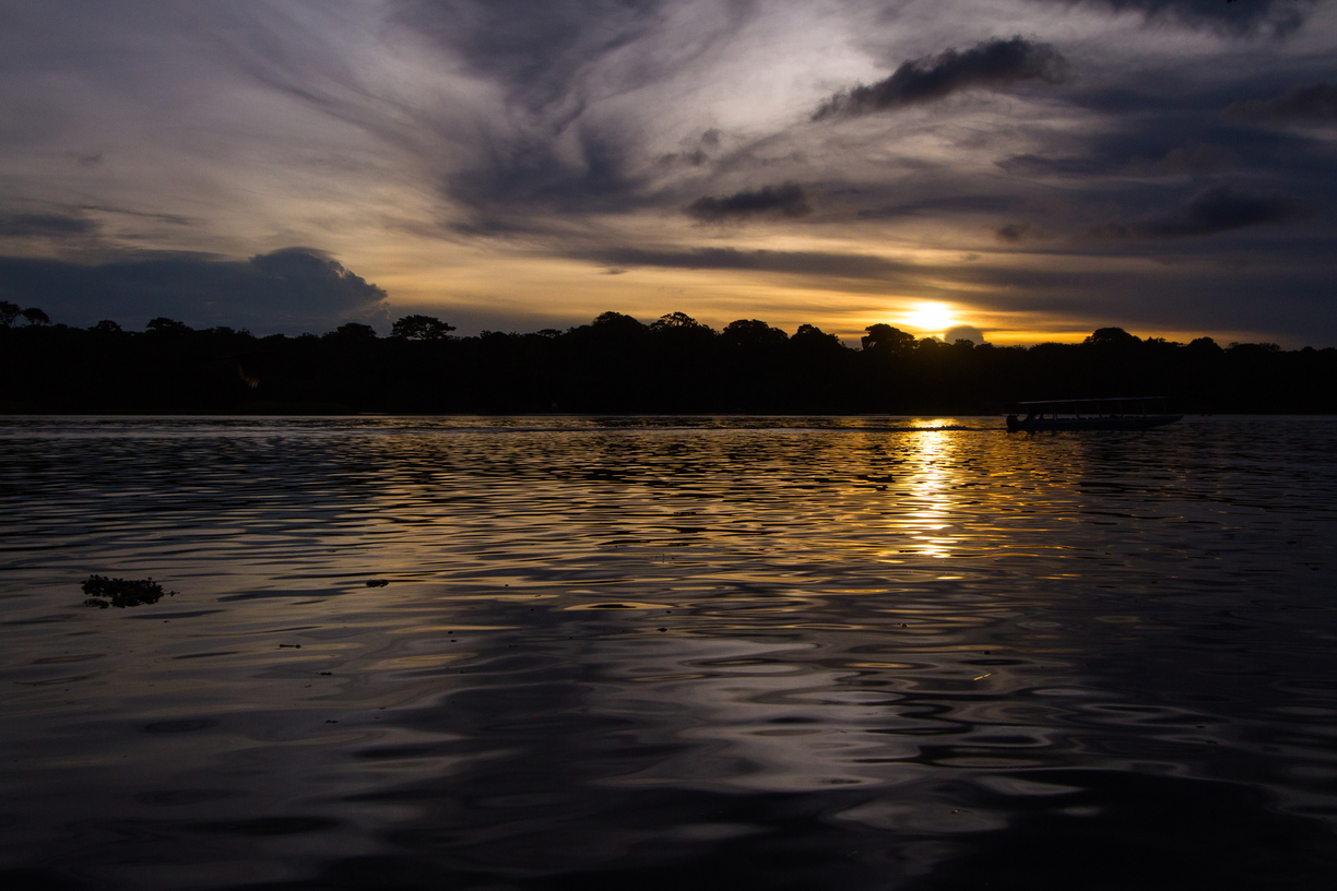 As the sun sets, an empty river boat returns to dock on the narrow spit of land that is Tortuguero, Costa Rica, closely followed by a barely visible silhouetted bird in flight.
